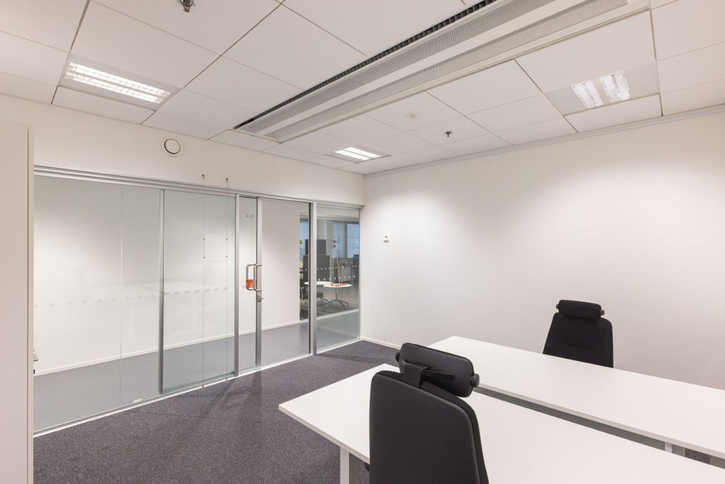 Office space with two desks and two office chairs. One wall is glass and features a glass sliding door to the corridor.
