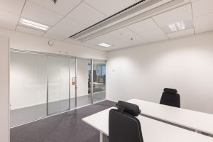 Office space with two desks and two office chairs. One wall is glass and features a glass sliding door to the corridor.