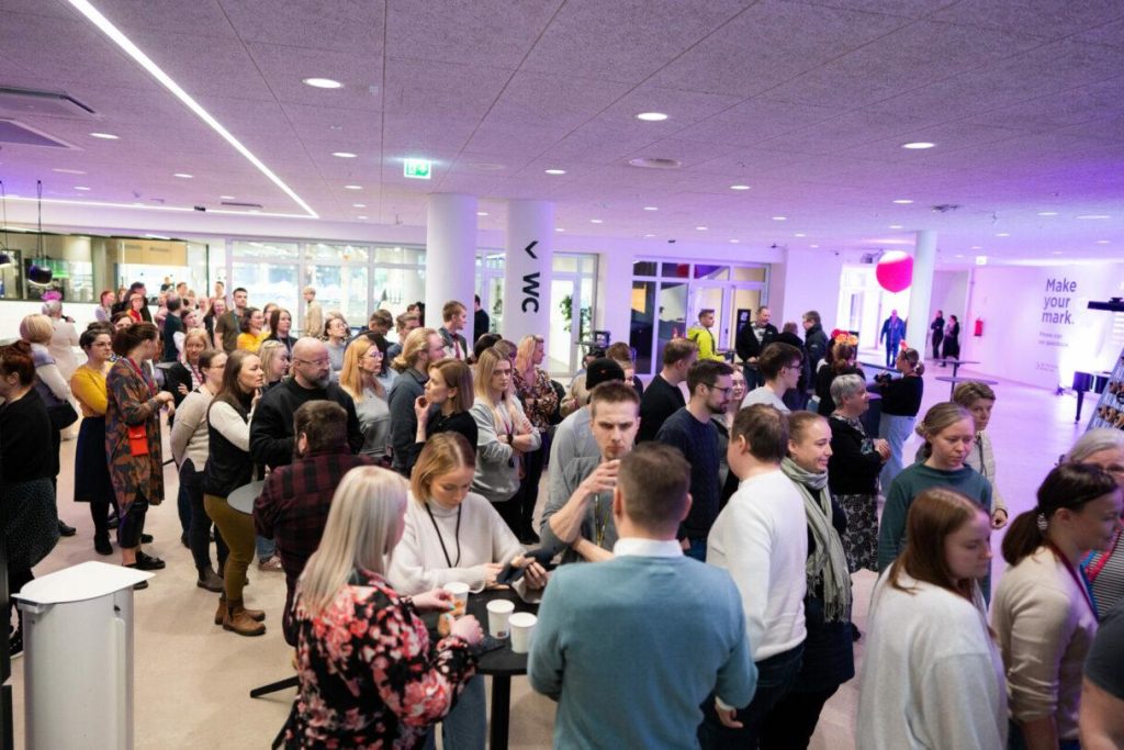 Suuri yleisö kokoontuneena tapahtumatilan aulaan korkeiden pöytien ympärille, taustalla esitysnäyttö ja opasteita.