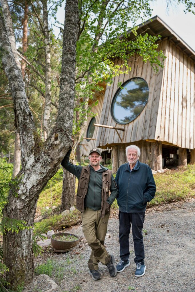 Kaksi henkilöä seisoo metsäisessä ympäristössä puisen rakennuksen edustalla, taustalla pyöreäikkunainen puurakennus.