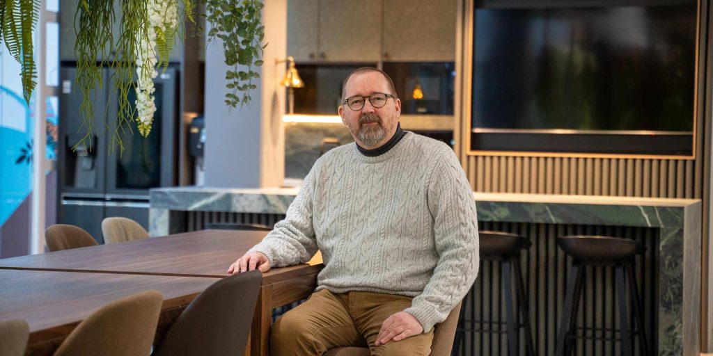 Henkilö istuu suuren pöydän äärellä toimistotilan keittiö- ja oleskelualueella, taustalla näyttö ja baarijakkaroita.