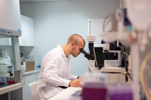 Henkilö tarkastelee näytettä mikroskoopilla laboratoriossa, ympärillä tutkimuslaitteita ja työpisteitä.