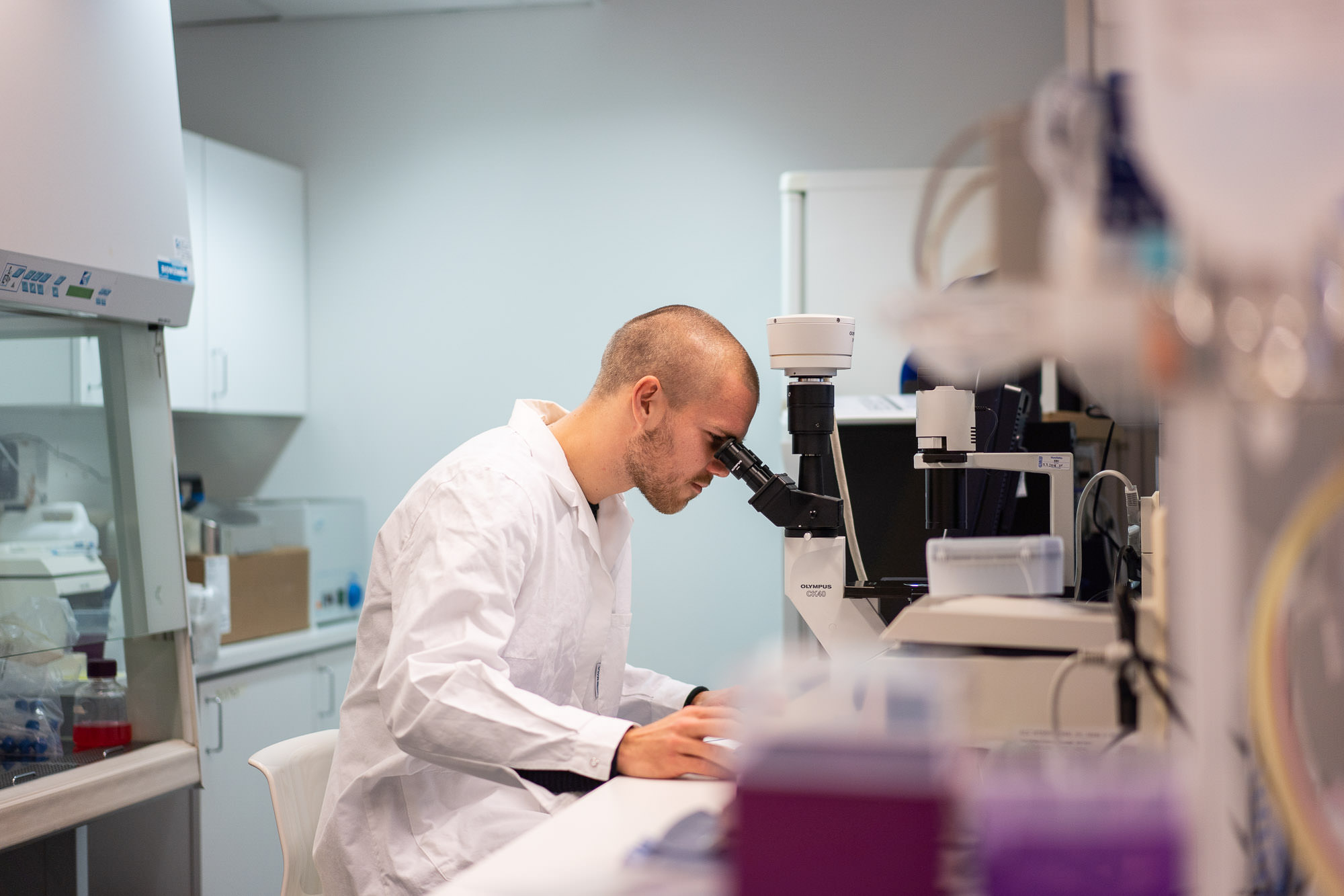 Henkilö tarkastelee näytettä mikroskoopilla laboratoriossa, ympärillä tutkimuslaitteita ja työpisteitä.