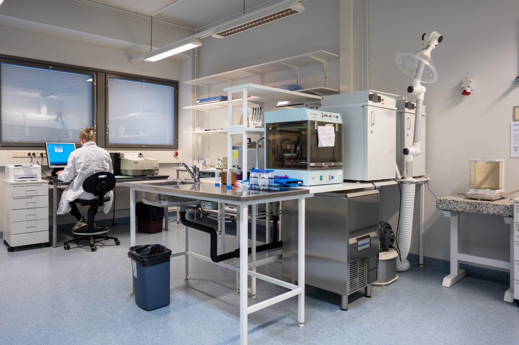 Laaja laboratoriotila, jossa työpöytiä, tutkimuslaitteita ja yksi henkilö työskentelemässä tietokoneen äärellä.