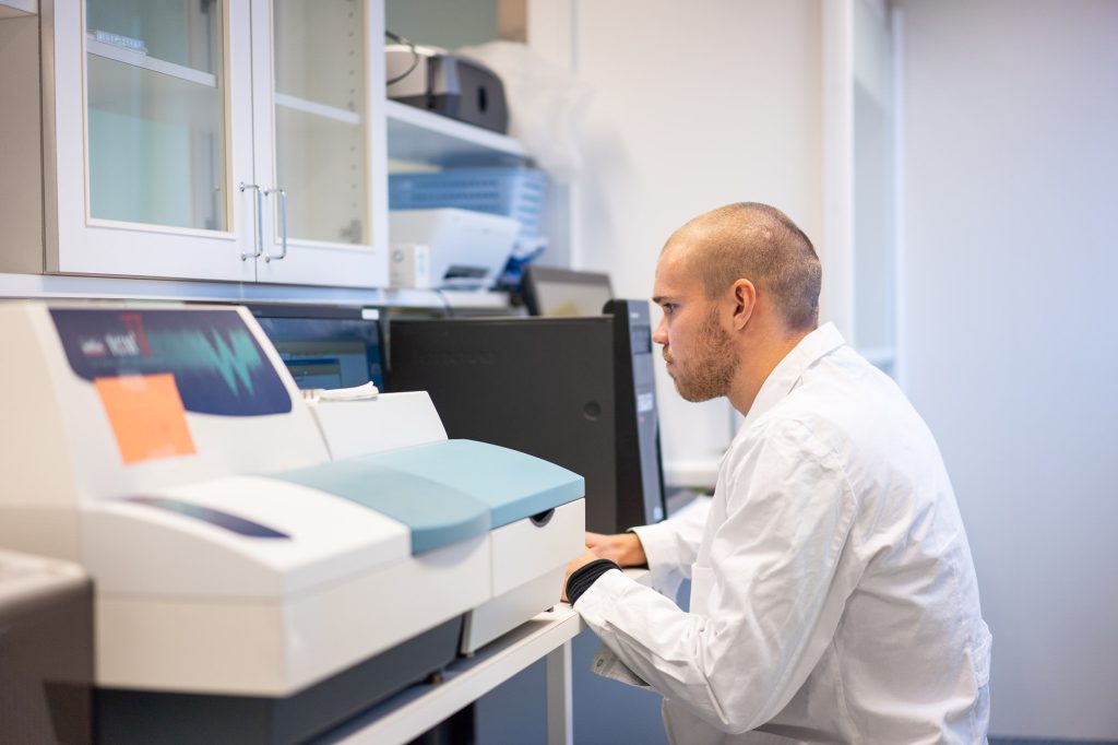 Henkilö työskentelee laboratoriossa analyysilaitteen äärellä, ympärillä kaappeja ja tietokoneita.