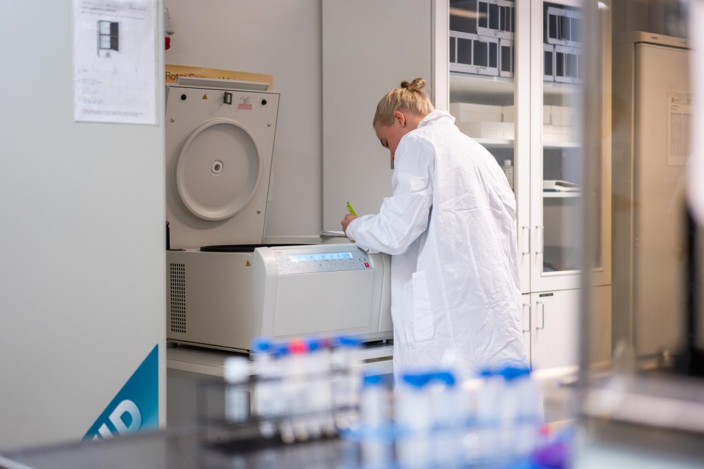 Henkilö työskentelee laboratoriossa sentrifugin äärellä ja tekee muistiinpanoja, ympärillä kaappeja ja työpintoja.