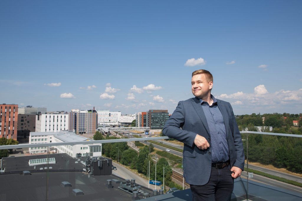 Henkilö seisoo kattoterassilla kaiteen äärellä, taustalla kaupunkimaisema ja rakennuksia aurinkoisena päivänä.