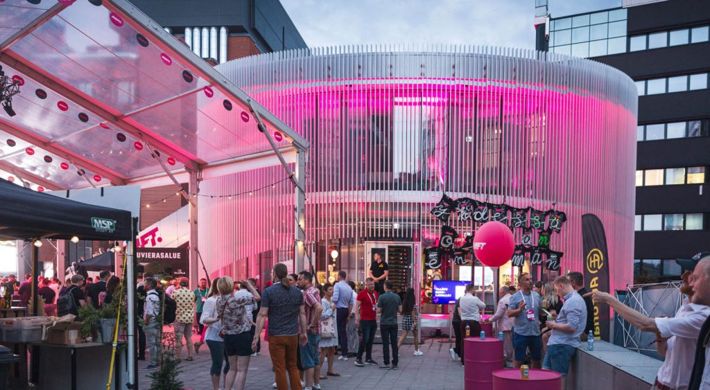 Ihmiset keskustelevat ja liikkuvat tapahtuma-alueella SHIFT businessfestivaalilla, taustalla valaistu pyöreä rakennus ja tapahtumarakenteita.