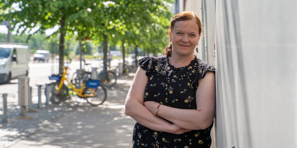 Henkilö seisoo ulkona katukäytävällä rakennuksen seinää vasten, taustalla puita, polkupyöriä ja kaupunkiympäristö.