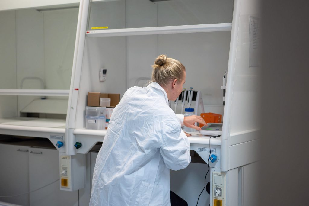 Henkilö työskentelee vetokaapissa laboratoriossa, edessä mittauslaite ja laboratorion perusvälineitä.