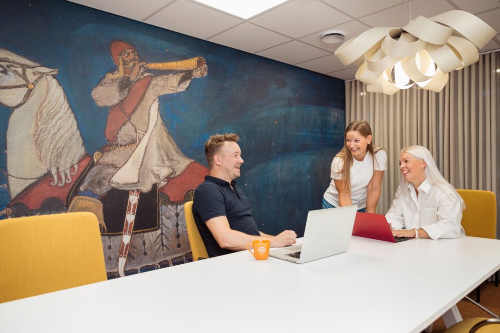 Neuvottelutila, jossa useita ihmisiä istuu pöydän äärellä kannettavien tietokoneiden kanssa, taustalla seinämaalaus.