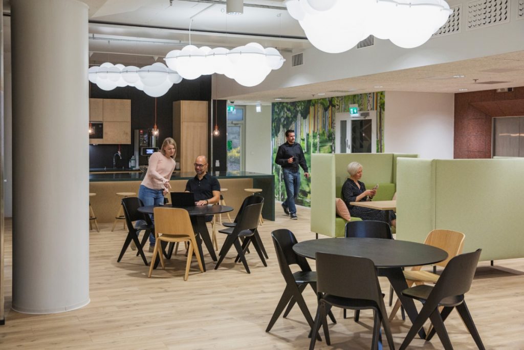 Ihmiset työskentelevät ja keskustelevat Werstaa coworking‑tilassa, jossa on pilvimuotoiset valaisimet, keittiötausta ja vihreitä työskentelykoppeja.