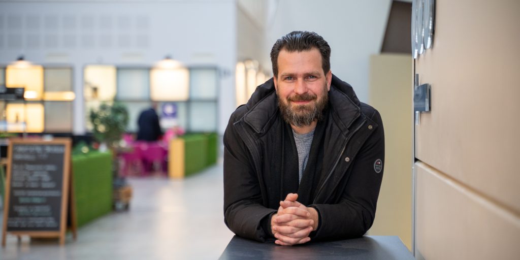 Henkilö nojaa pöytään aulatilassa, taustalla kahvilamainen ympäristö ja istuinryhmiä.