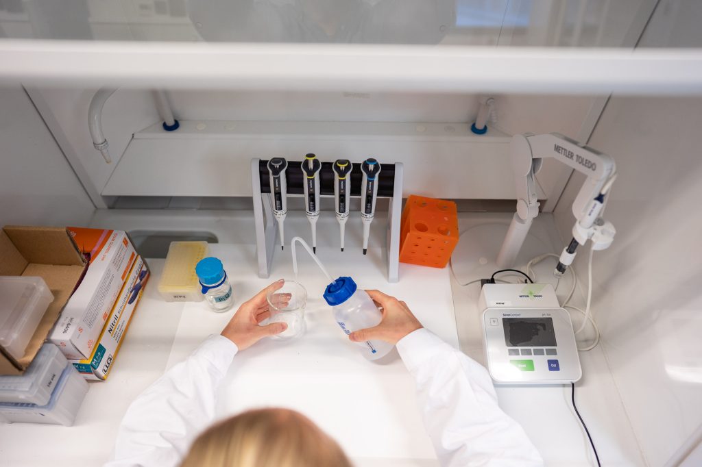 Lähikuva laboratoriotyöpöydästä ylhäältä päin, käsissä mitta-astia ja pullo, taustalla pipetit ja mittauslaite.