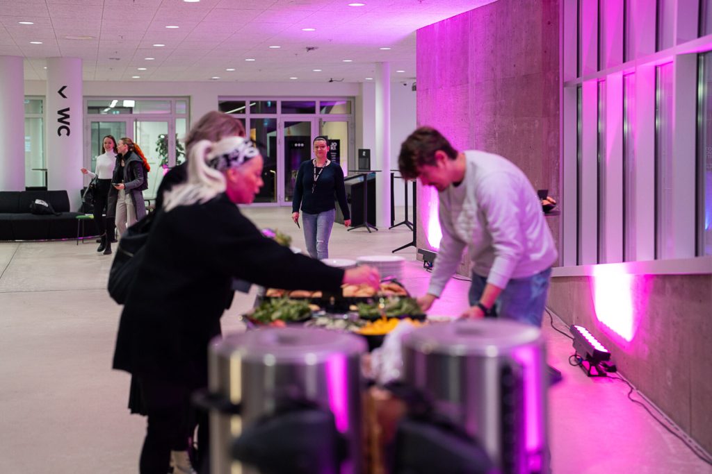 Ihmiset kokoavat tarjoiluja tapahtumatilassa, etualalla buffetpöytä ja taustalla avara aula violetilla valaistuksella.