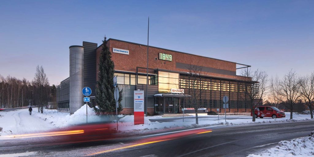 Tiilirakenteinen rakennus talvimaisemassa iltahämärässä, valaistus näkyy ikkunoista ja kadulla kulkee autoja.