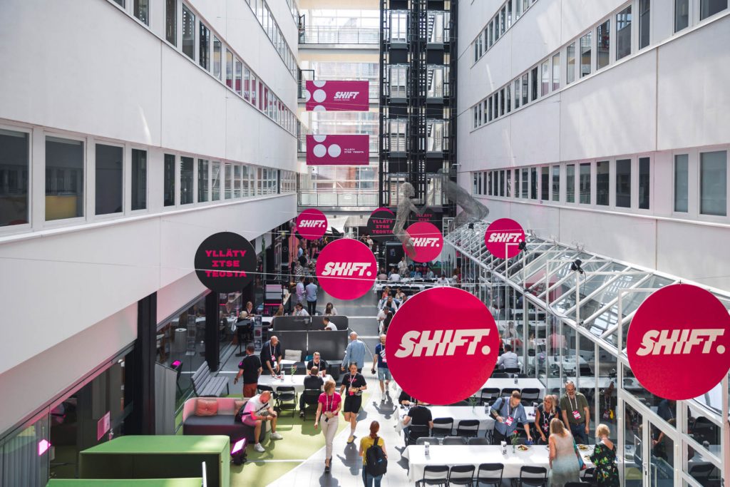 SHIFT businessfestivaalin sisäpiha, jossa ihmiset työskentelevät ja keskustelevat pöytien äärellä, yläpuolella roikkuvat SHIFT‑logot.