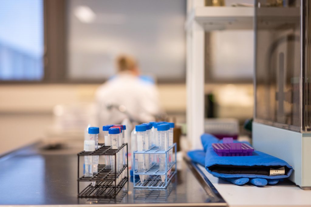 Koeputkia telineissä laboratoriotyöpöydällä, vieressä suojakäsineitä ja tarvikkeita, taustalla työskentelevä henkilö epätarkkana.