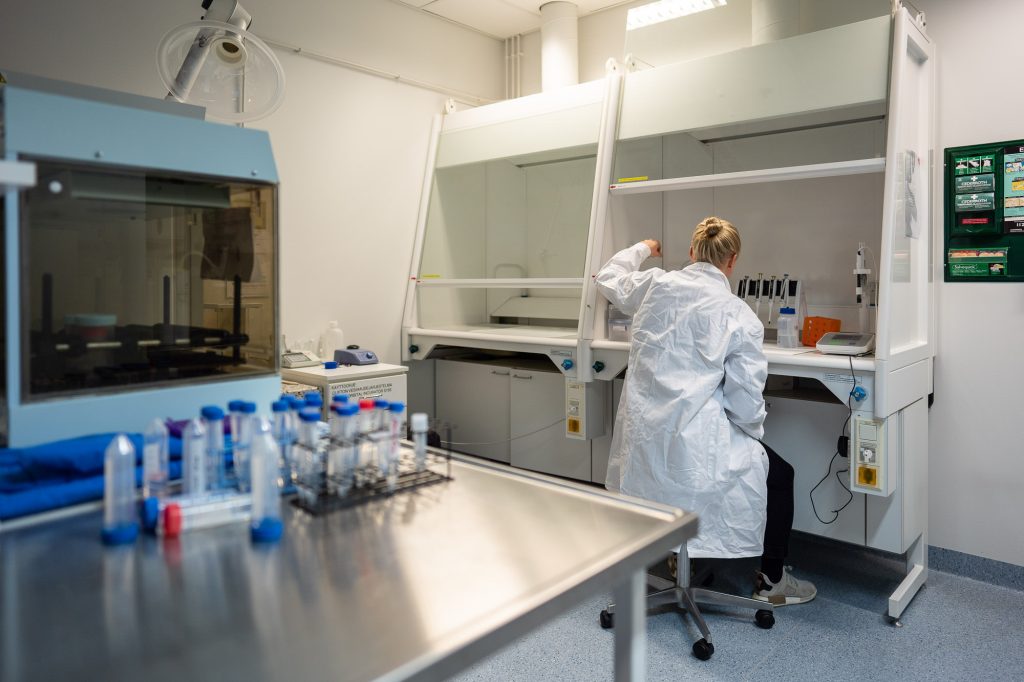 Henkilö työskentelee vetokaapissa laboratoriossa, edessä pipettejä ja mittausvälineitä, ympärillä laboratorion peruskalusteita.