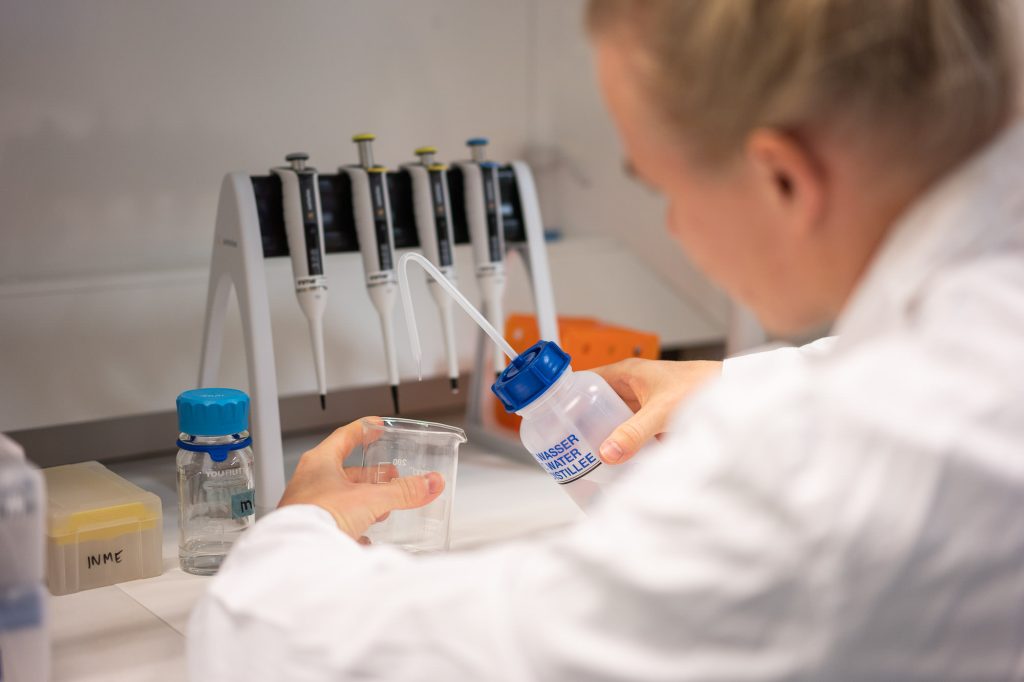 Henkilö kaataa nestettä mitta-astiaan laboratoriotyöpisteellä, taustalla pipettejä ja näytepulloja.