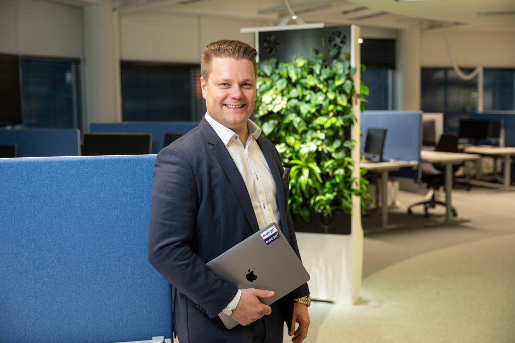 Henkilö seisoo toimistotilassa työpisteiden vieressä ja pitää kädessään kannettavaa tietokonetta, taustalla viherseinä.