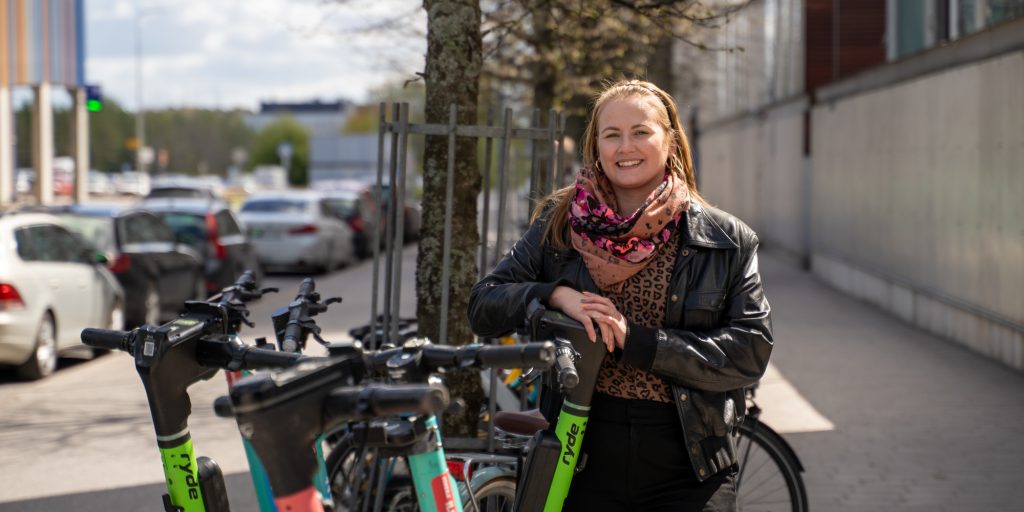 Henkilö seisoo ulkona pyöräpysäköinnin vieressä katutilassa, taustalla pysäköityjä autoja ja rakennuksia.