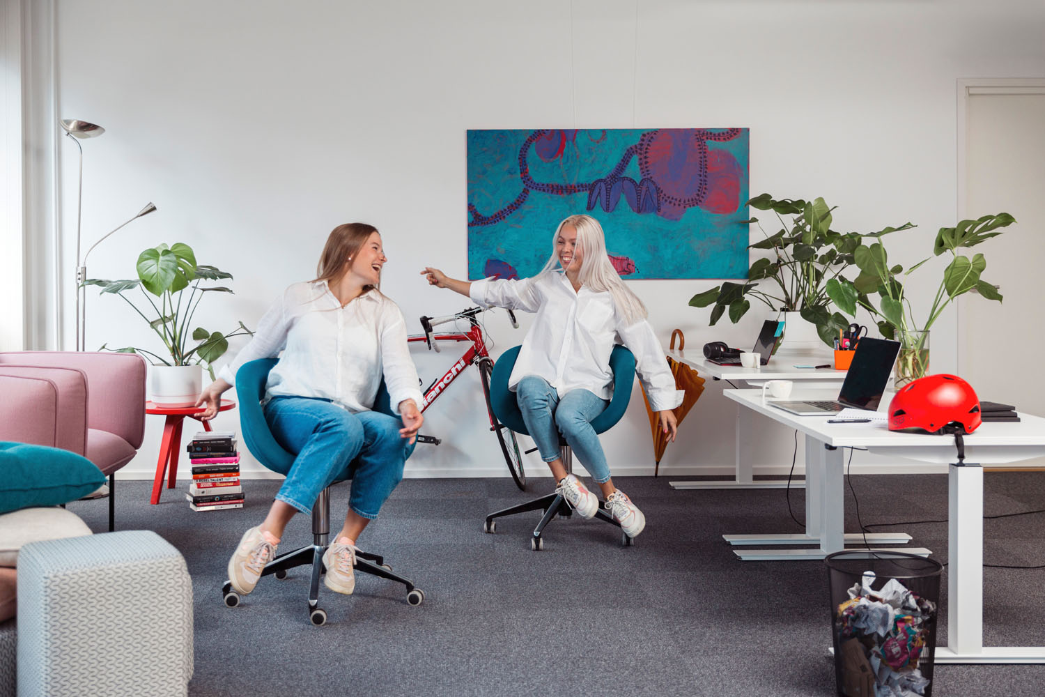 Kaksi naista pyörii tuoleilla toimistossa, jossa on sähköpöydät, sohva, viherkasveja ja pyörä. 