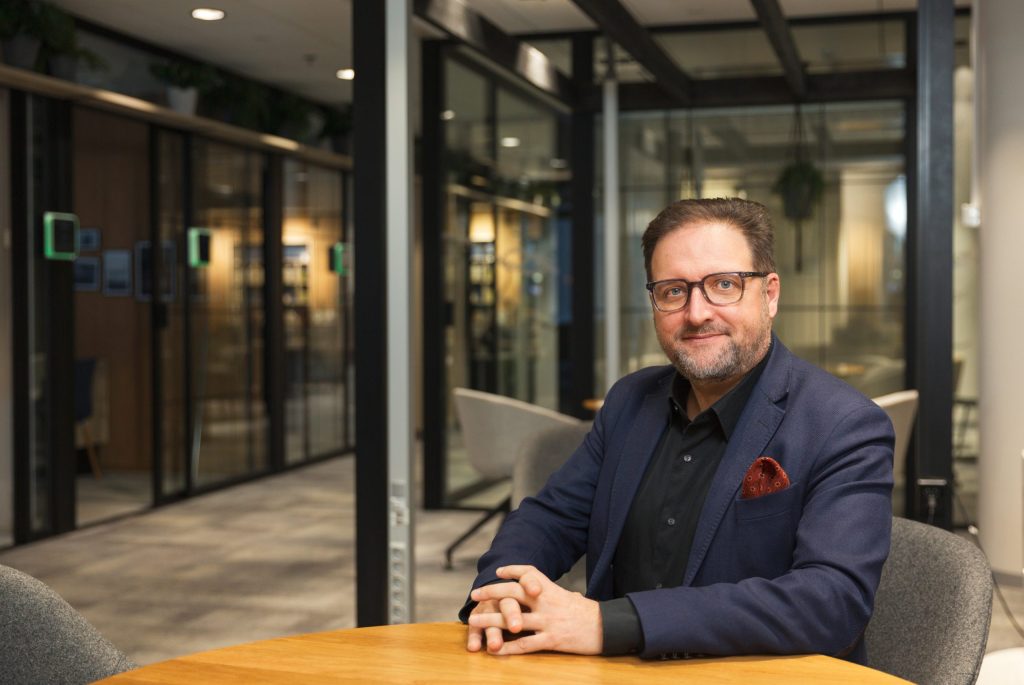 Henkilö istuu pöydän ääressä neuvottelutilassa, taustalla lasiseinäisiä huoneita ja moderni sisustus.