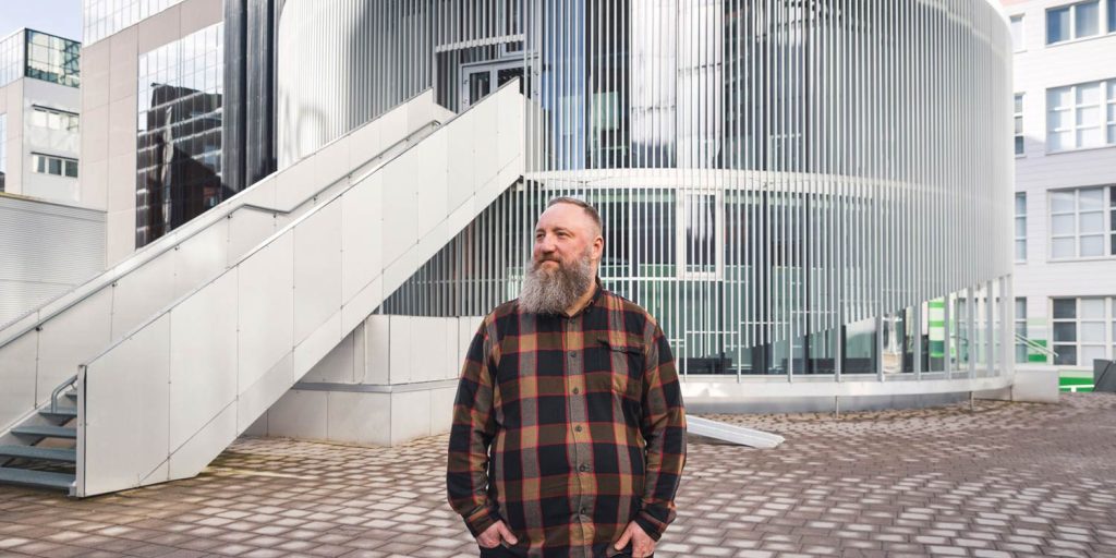 Henkilö seisoo ulkotilassa modernin, pyöreälinjaisen rakennuksen edessä, taustalla portaikko ja metallinen julkisivu.