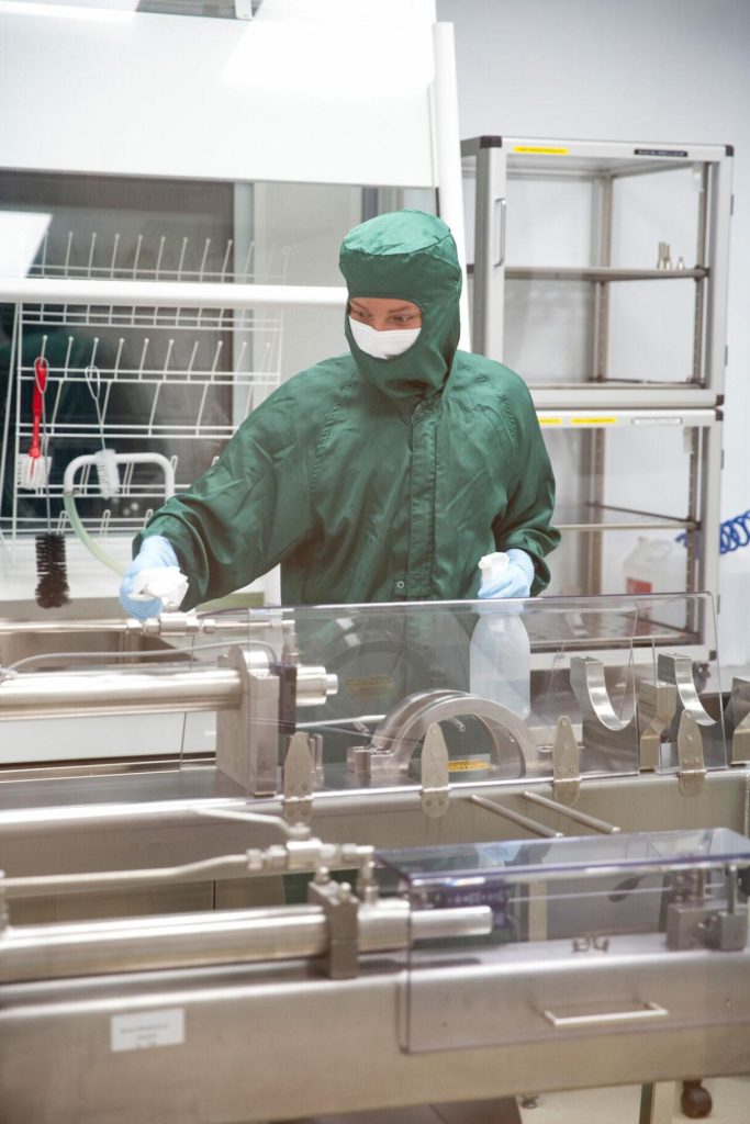 Henkilö työskentelee suojavaatetuksessa tuotanto- tai laboratoriotilassa, edessä teollinen laitteisto ja läpinäkyvä suojaseinä.