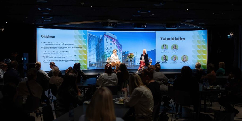 Paneelikeskustelu tapahtumatilassa, lavalla kaksi keskustelijaa ja yleisö seuraa esitystä pöytien äärellä.