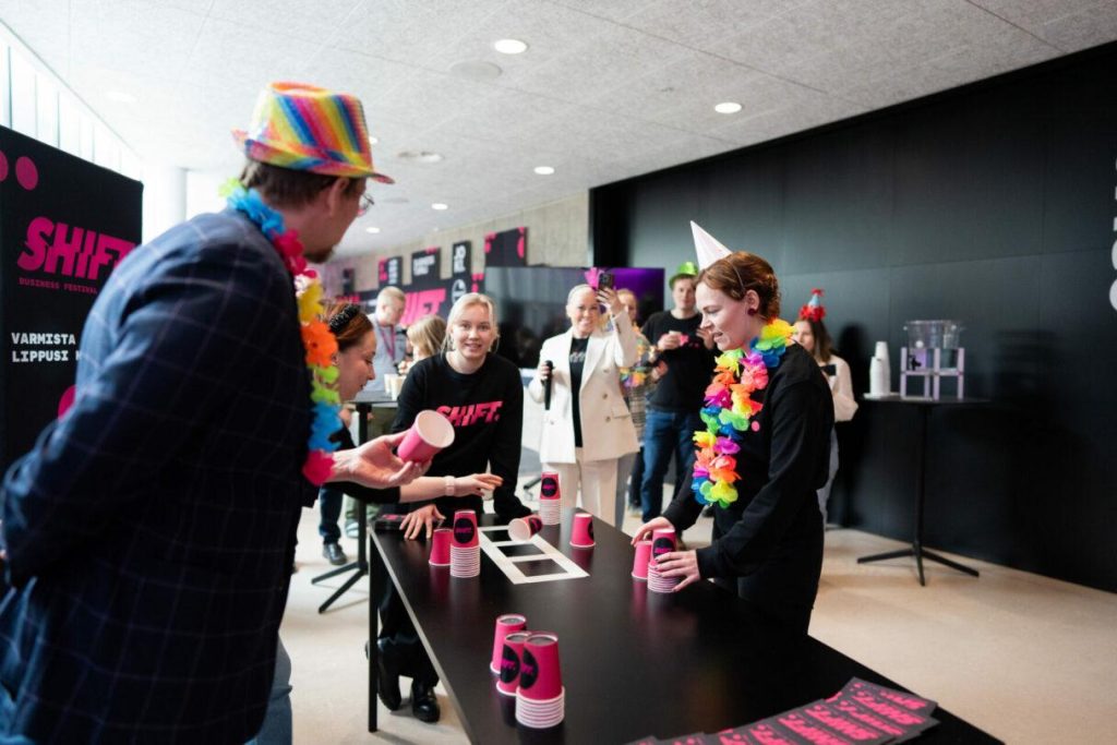 Tapahtumapiste, jossa vieraat osallistuvat pöytäpelimäiseen aktiviteettiin, pöydällä pinkkejä juomakuppeja ja taustalla tapahtumabrändäys.