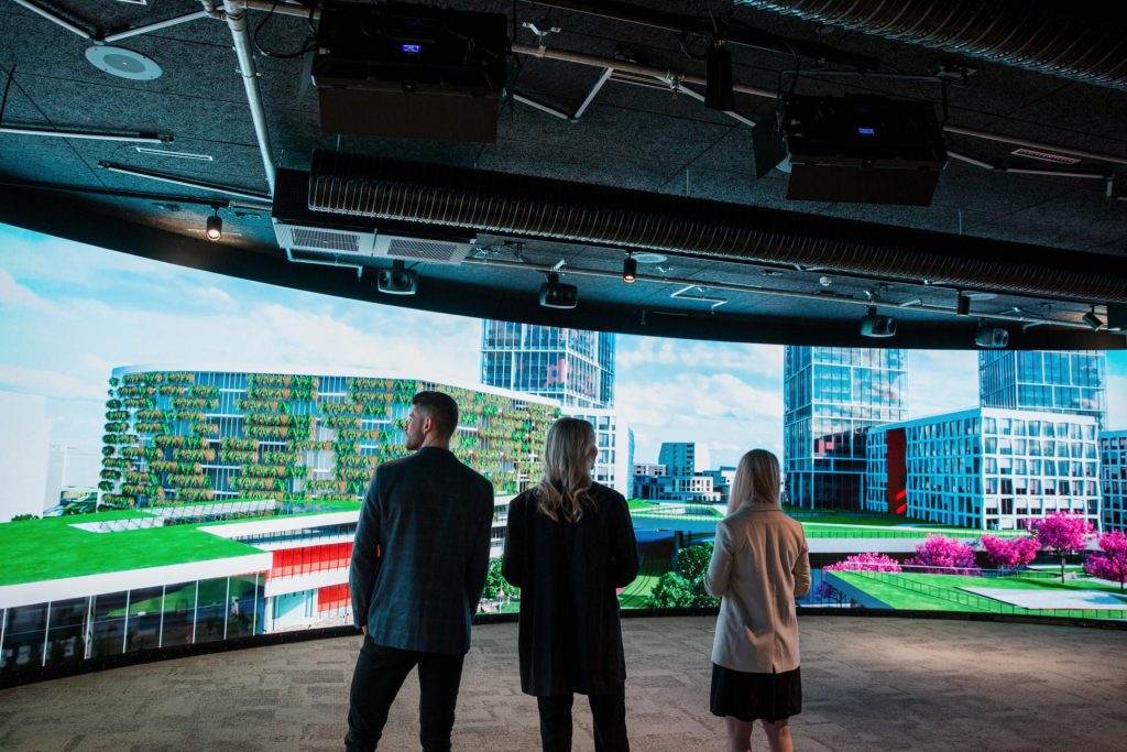 Kolme henkilöä seisoo suuren LED-näytön edessä, jossa esitetään havainnekuvaa kaupunkiympäristöstä.
