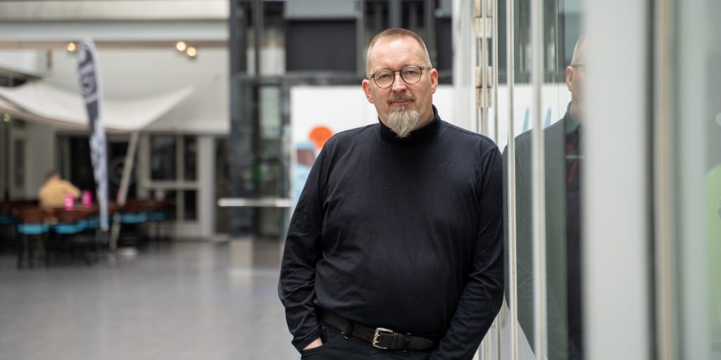 Henkilö nojaa lasiseinään aulatilassa, taustalla avara sisätila ja istuinryhmiä.
