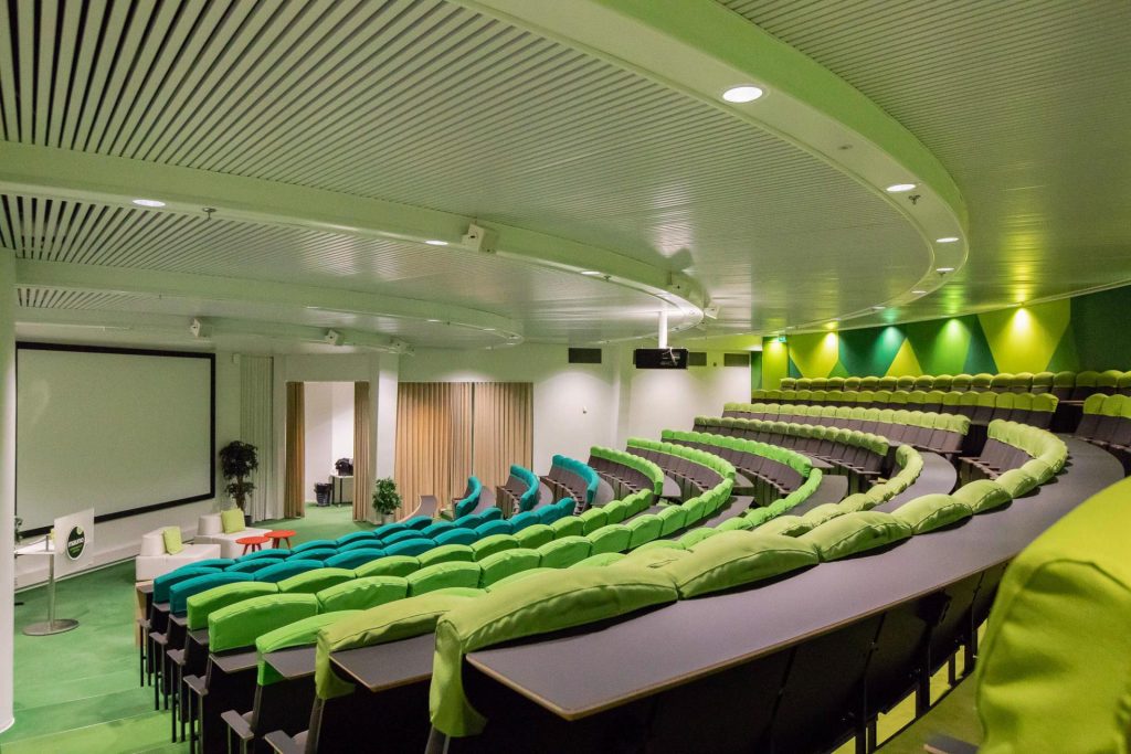 Luokkamuotoinen auditorio, jossa on vihreitä istuimia, esitysnäyttö ja valaistu tila tapahtumia ja luentoja varten.