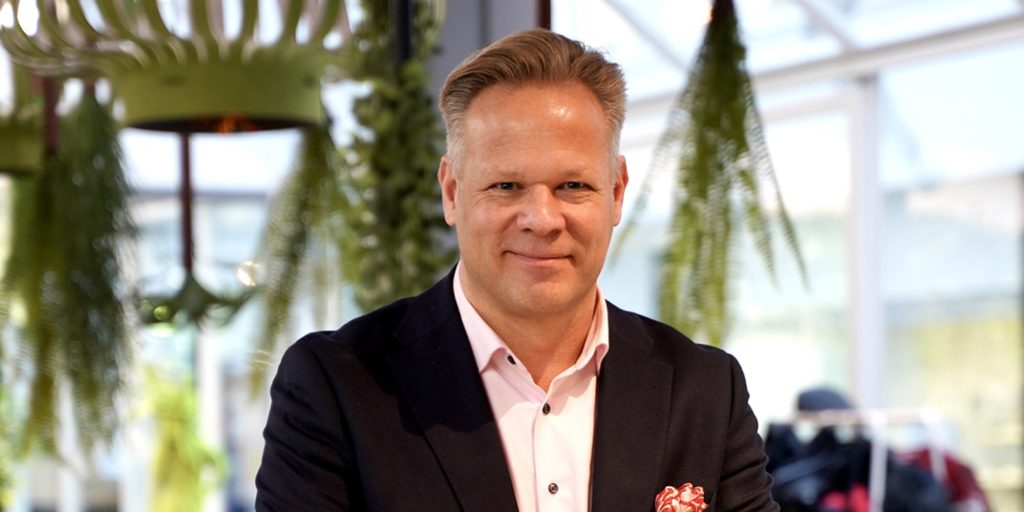 Henkilö seisoo tukeutuen tasoa vasten, takanaan suuri pöytä, jonka ääressä tuoleja ja yläpuolella huonekasveja
