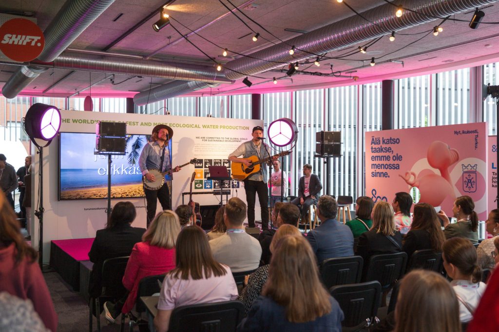 Bändi esiintyy sisälavalla SHIFT businessfestivaalilla, yleisö istuu riveissä lavan edessä ja seuraa esitystä.