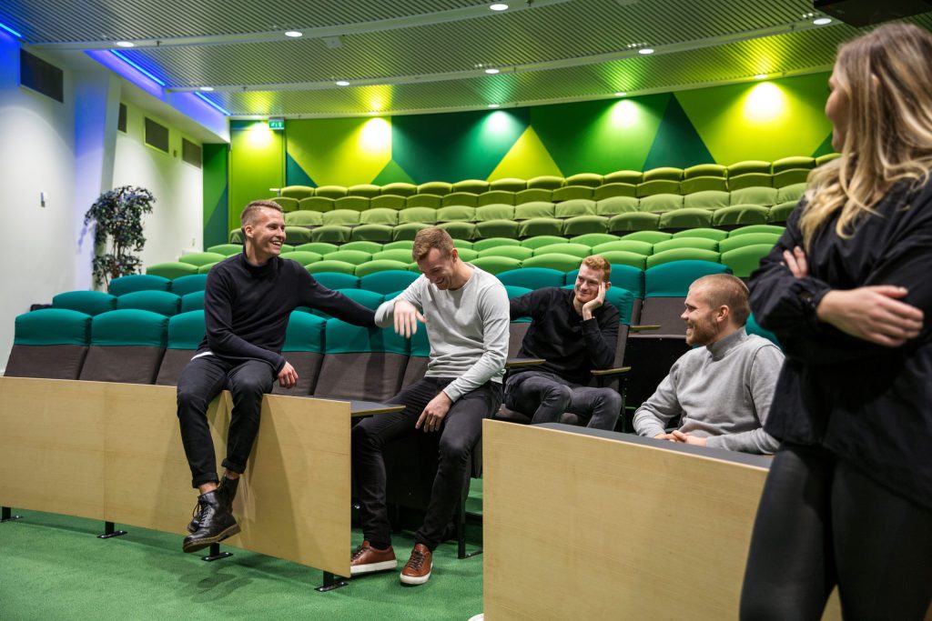 Ryhmä ihmisiä keskustelee ja istuu auditoriomaisessa tilassa, jossa on vihreät katsomoistuimet ja valaistu sisustus.